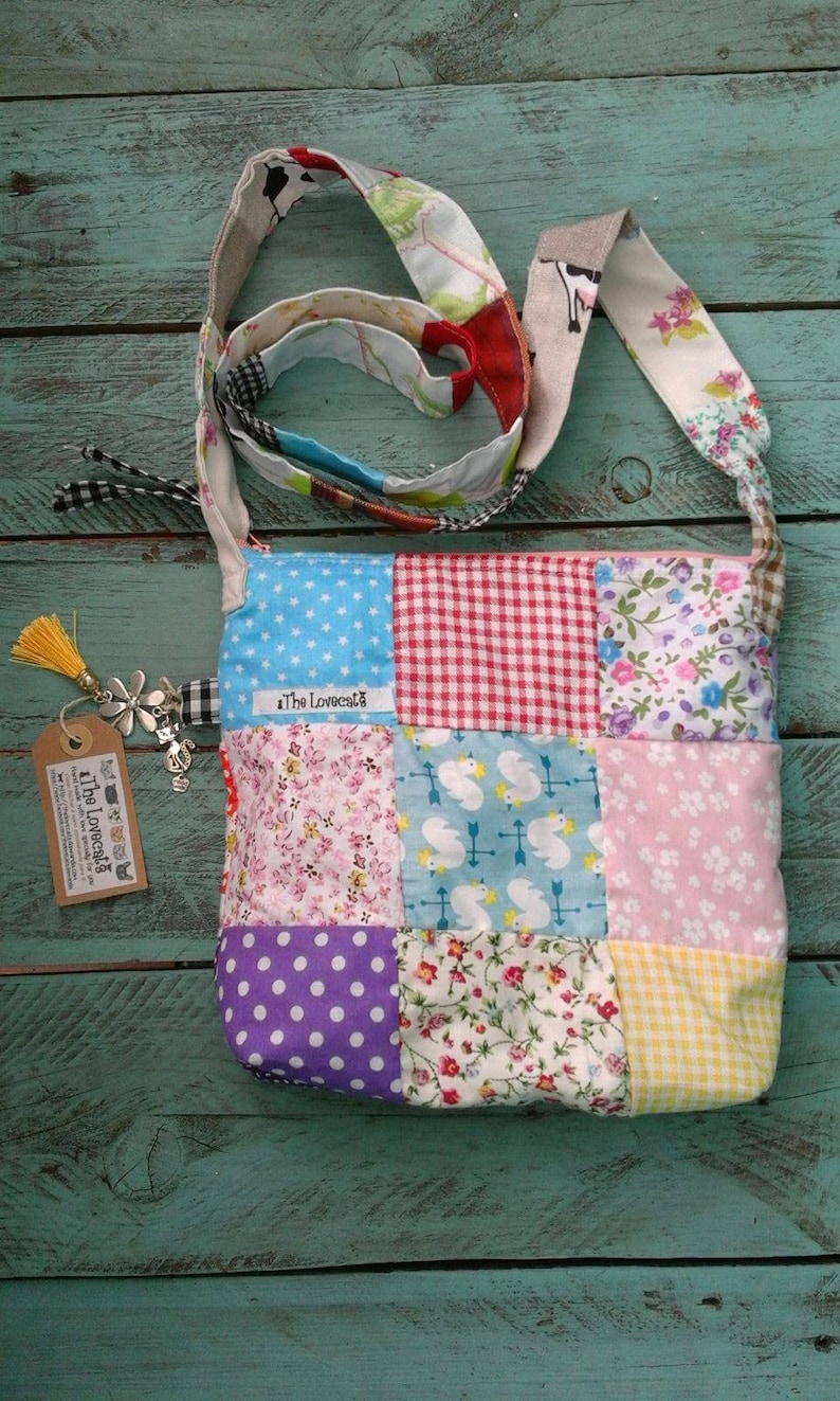 May include: A patchwork crossbody bag with a variety of fabric patterns, including polka dots, gingham, and floral prints. The bag has a long strap and a tag that says "The Lovecats". The bag is made of various colors, including blue, pink, red, and yellow.