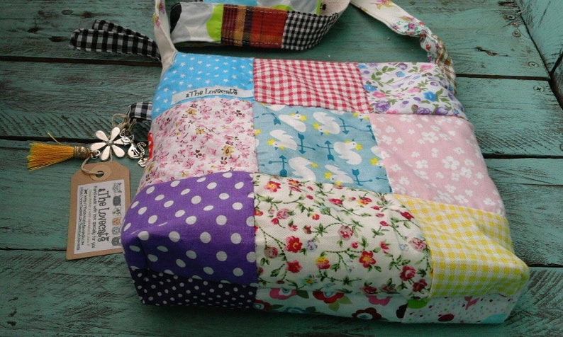 May include: A patchwork shoulder bag with a variety of fabric patterns, including polka dots, floral prints, and gingham. The bag has a matching strap and a tag with the text "The Lovecats". A decorative tassel and charm are attached.