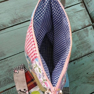 May include: Open view of a patchwork fabric bag with a pink zipper. The interior is lined with blue and white gingham fabric. Exterior patches include floral, gingham, and plaid patterns. A fabric pull extends from the zipper.