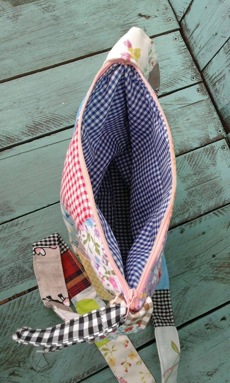 May include: Open view of a patchwork fabric bag with a pink zipper. The interior is lined with blue and white gingham fabric. Exterior patches include floral, gingham, and plaid patterns. A fabric pull extends from the zipper.