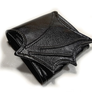 May include: A black leather wallet shaped like a bat wing. The wallet is folded closed and the bat wing design is prominent.
