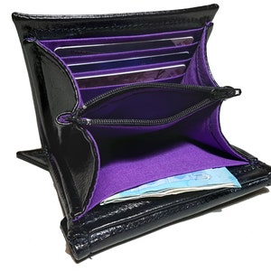 May include: Black leather wallet with a purple interior. The wallet has multiple card slots and a zippered compartment. The wallet is open and there is a bill inside.