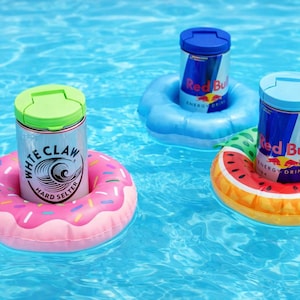 Puede incluir: Tres latas de bebidas flotando en una piscina sobre flotadores inflables. Una lata de White Claw Hard Seltzer en un flotador de rosquilla rosa. Dos latas de Red Bull flotan sobre flotadores azules y con forma de sand&iacute;a.