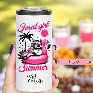 May include: White insulated can cooler with a pink flamingo float and a raccoon wearing sunglasses. The text on the cooler reads "Feral girl summer Mia" and "fits slim cans".