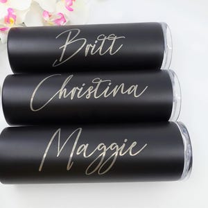 Può includere: Tre bicchieri termici neri con coperchi trasparenti sono impilati, ognuno con un nome in elegante corsivo: Britt, Christina e Maggie. Orchidee bianche e rosa sullo sfondo.