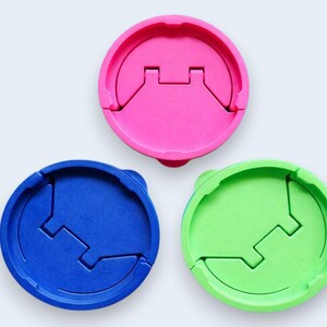 Puede incluir: Tres tapas redondas de pl&aacute;stico de colores: rosa, azul y verde. Cada tapa tiene un dise&ntilde;o &uacute;nico con un recorte rectangular central y una secci&oacute;n con bisagras. Las tapas est&aacute;n dispuestas sobre un fondo blanco.