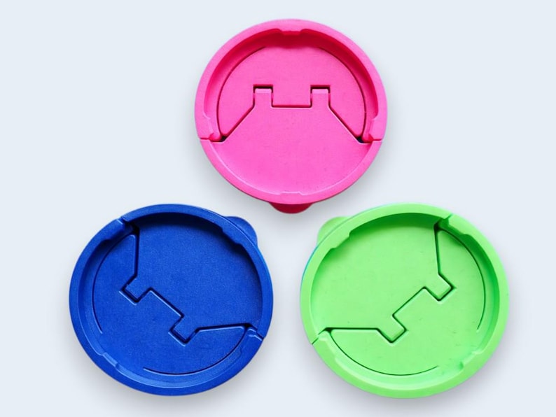 Puede incluir: Tres tapas redondas de pl&aacute;stico de colores: rosa, azul y verde. Cada tapa tiene un dise&ntilde;o &uacute;nico con un recorte rectangular central y una secci&oacute;n con bisagras. Las tapas est&aacute;n dispuestas sobre un fondo blanco.
