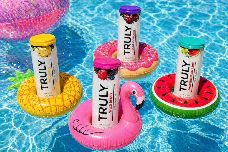 Puede incluir: Cuatro latas de Truly Hard Seltzer flotando en una piscina, cada una en un portavasos inflable diferente. Los sabores incluyen Pi&ntilde;a, Cereza Negra, Baya Silvestre y Sand&iacute;a Lima. Los portavasos tienen forma de pi&ntilde;a, donut, flamenco y rodaja de sand&iacute;a.