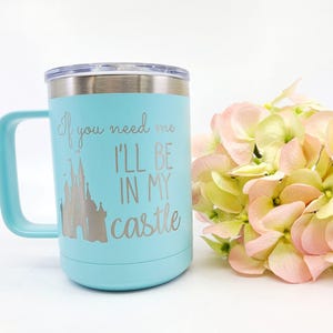 May include: A light blue travel mug with a silver lid and handle. The mug features the text "If you need me I'll be in my castle" with a castle graphic. A bouquet of pink and yellow flowers is next to the mug.
