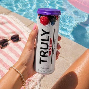 Puede incluir: Una lata de Truly Wild Berry Hard Seltzer con tapa morada y dise&ntilde;o de bayas. La lata es sostenida por una persona cerca de una piscina. La lata dice "TRULY WILD BERRY HARD SELTZER" y "5% ALC./VOL. 355ml".