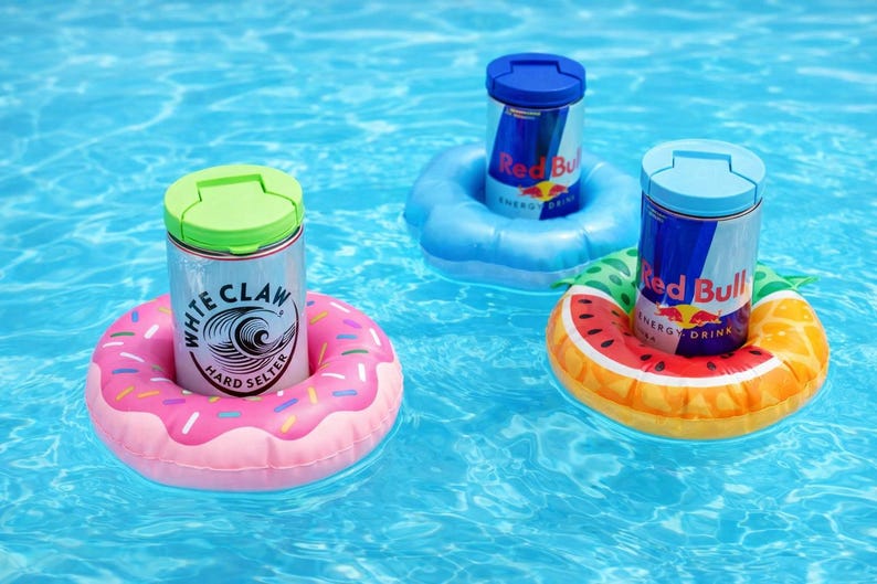 Puede incluir: Tres latas de bebidas flotando en una piscina sobre flotadores inflables. Una lata de White Claw Hard Seltzer en un flotador de rosquilla rosa. Dos latas de Red Bull flotan sobre flotadores azules y con forma de sand&iacute;a.