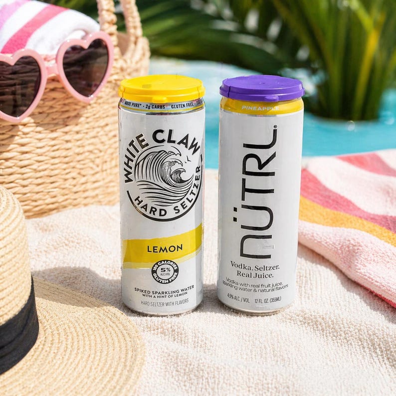Puede incluir: Dos latas de hard seltzer sobre una toalla de playa. Una lata est&aacute; etiquetada como "White Claw Lemon" con tapa amarilla. La otra lata est&aacute; etiquetada como "N&Uuml;TRL Pineapple" con tapa morada. Ambas latas son blancas con texto negro.