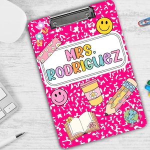 Puede incluir: Portapapeles rosa con un dise&ntilde;o estampado blanco y negro. El portapapeles tiene una funda de pl&aacute;stico transparente y un clip de metal. El portapapeles tiene una pegatina blanca con el texto "Mrs. Rodriguez" en letras de arco&iacute;ris. El portapapeles tambi&eacute;n tiene varias pegatinas, incluyendo una cara sonriente, un l&aacute;piz, una taza de caf&eacute;, un libro y un globo terr&aacute;queo.