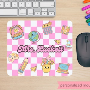 May include: A personalized mouse pad with a pink and white checkered pattern. The design includes school-themed illustrations like a backpack, globe, calculator, and pencil. The name "Mrs. Luckett" is displayed in a retro font.