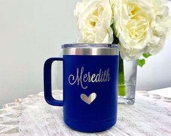 May include: A navy blue insulated coffee mug with a silver rim and handle. The mug is engraved with the name "Meredith" in cursive and a silver heart. A bouquet of white roses sits in the background.