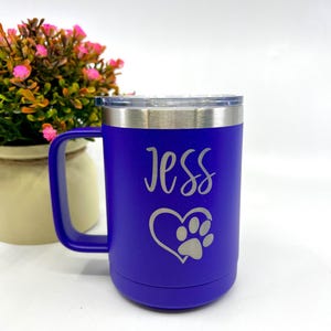 May include: A deep blue travel mug with a silver rim and handle. The mug features the name "Jess" in cursive, along with a heart and paw print design. The mug is shown next to a small pot of pink flowers.