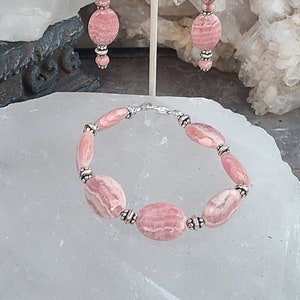 May include: A pink and silver bracelet and earrings set made with oval-shaped pink stones. The bracelet has a silver clasp and the earrings have silver hooks.