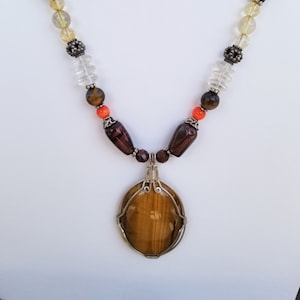 Puede incluir: Un collar de piedras preciosas doradas y marrones con un colgante de ojo de tigre. El collar presenta una variedad de cuentas, incluyendo cuentas transparentes, naranjas y marrones.
