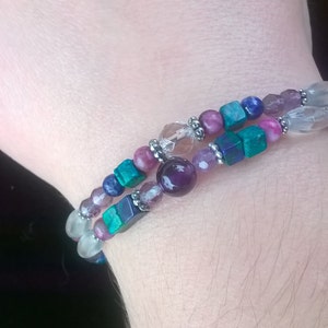 May include: A multi-strand bracelet featuring beads of various colours and shapes, including purple, pink, blue, green, and white. The bracelet is made from natural stones and has a silver charm.