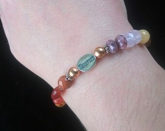Puede incluir: Un brazalete de cuentas multicolores con un dije de plata que dice "Taurus".
