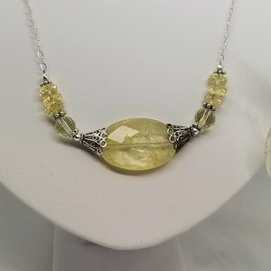 Puede incluir: Un collar de cadena de plata con un gran colgante ovalado de piedra preciosa de color amarillo claro. El colgante est&aacute; rodeado de detalles plateados y la cadena est&aacute; adornada con cuentas m&aacute;s peque&ntilde;as de color amarillo claro.