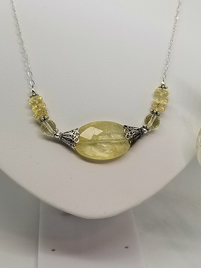 Puede incluir: Un collar de cadena de plata con un gran colgante ovalado de piedra preciosa de color amarillo claro. El colgante est&aacute; rodeado de detalles plateados y la cadena est&aacute; adornada con cuentas m&aacute;s peque&ntilde;as de color amarillo claro.