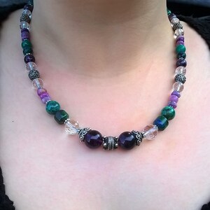 May include: A multi-coloured gemstone necklace with green, purple and clear beads. The necklace has two larger purple beads with silver accents in the centre.