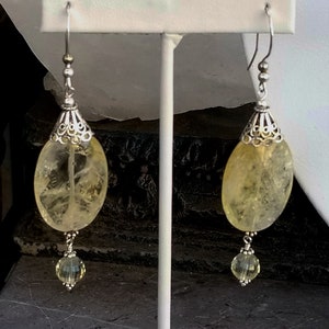 Puede incluir: Un par de pendientes de plata con piedras de citrino amarillo y detalles de cristal transparente. Los pendientes est&aacute;n colgados de un expositor blanco.