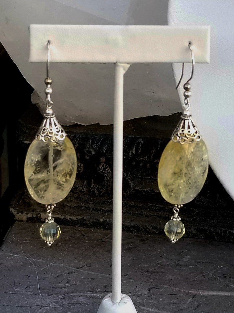 Puede incluir: Un par de pendientes de plata con piedras de citrino amarillo y detalles de cristal transparente. Los pendientes est&aacute;n colgados de un expositor blanco.