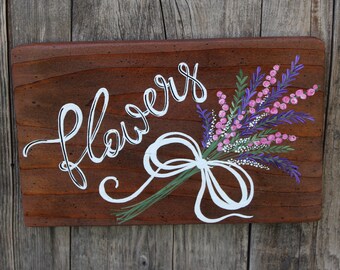 May include: A rectangular wooden sign with a dark brown stained finish featuring the word flowers painted in white script. A hand-painted bouquet of pink and purple flowers tied with a white ribbon is positioned to the right of the text.