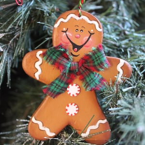 May include: A gingerbread man ornament featuring a brown body with white icing details on the head, arms, and legs. The figure has a painted face with rosy cheeks, two peppermint candy buttons, and a red and green plaid fabric bow tie.