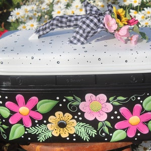 May include: A black and white painted storage box with a handle and a decorative bow. The box features a white lid with black polka dots and a black base adorned with pink and yellow flowers and green leaves. A floral arrangement sits on top.
