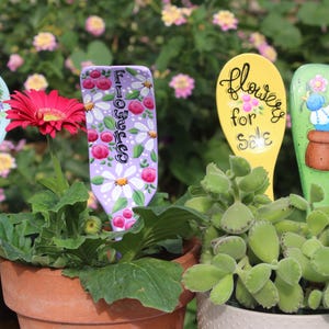 May include: A collection of hand-painted garden markers in various shapes and colors, including a light blue spoon with a heart cutout, a purple marker with the word "Flowers", and a yellow marker with the words "Flowers for Sale". The markers are placed in pots with plants and flowers.