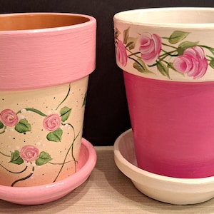 May include: Two ceramic flower pots with matching saucers. The left pot is cream-colored with pink roses and green vines, featuring a solid pink rim. The right pot is solid pink with a white rim decorated with a hand-painted pink rose and vine pattern.