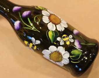 May include: A dark green glass bottle hand-painted with white daisies, yellow-centered flowers, purple thistles, and buzzing bees. The bottle's design includes green leaves and stems, creating a nature-inspired theme.