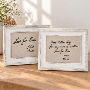 May include: Two rectangular picture frames with a distressed white finish and a beige fabric insert. The left frame features black embroidered text reading Love for Ever XXX Mum. The right frame features black embroidered text reading Happy Mothers day. from one mum to another Love for Ever XXX Mum.