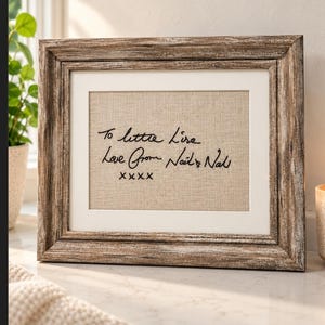 May include: A rectangular picture frame with a distressed brown wood finish containing a beige fabric insert. The insert features black embroidered cursive text that reads: To little Lise, Love from Nail & Nal, XXXX.