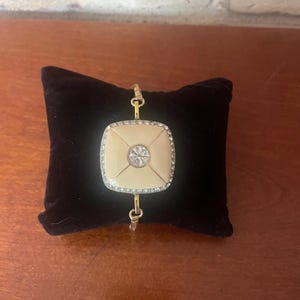 May include: Gold-toned bracelet featuring a cream-colored square centerpiece with a central clear stone and a border of smaller, sparkling stones. The bracelet is displayed on a black velvet cushion, showcasing its elegant design.