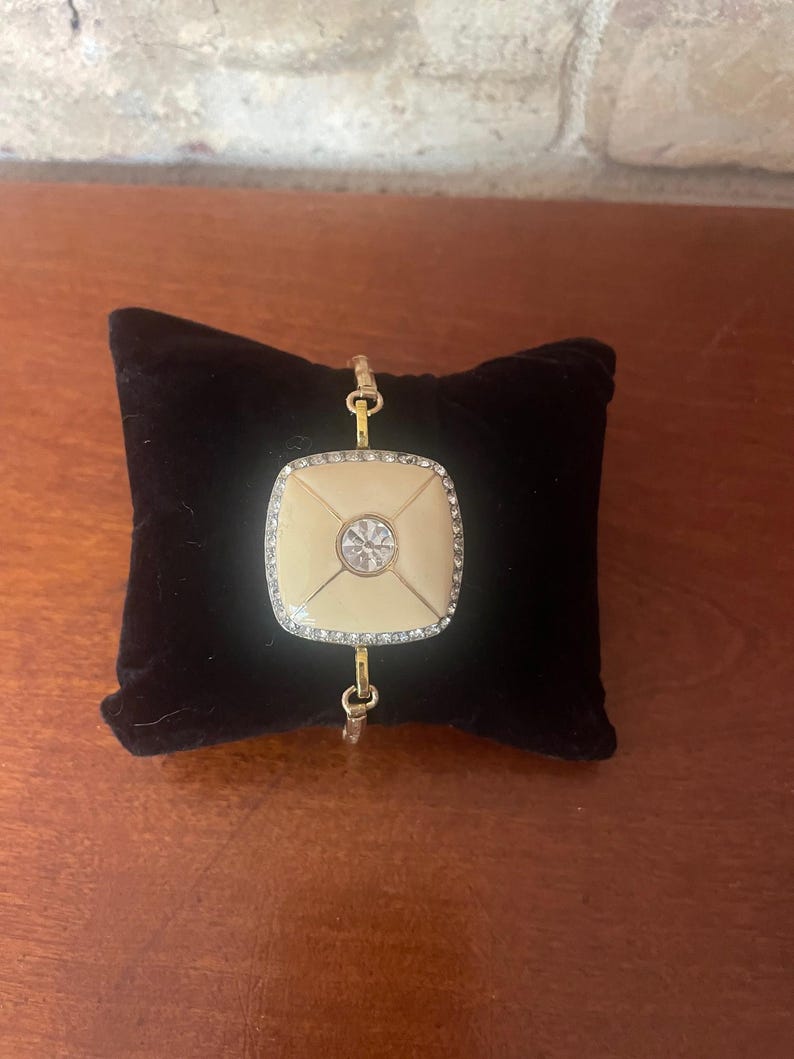 May include: Gold-toned bracelet featuring a cream-colored square centerpiece with a central clear stone and a border of smaller, sparkling stones. The bracelet is displayed on a black velvet cushion, showcasing its elegant design.