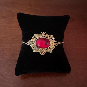 May include: A gold-toned bracelet featuring an oval-shaped, deep red gemstone centerpiece. The stone is set within an intricate, filigree-style metal frame with decorative scrollwork. The bracelet is attached to a thin, delicate gold-colored chain.