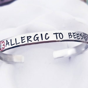 Può includere: Un braccialetto d'argento con il testo "ALLERGIC TO BEES" e un simbolo medico rosso e bianco. Un piccolo simbolo di alveare si trova sul lato destro del braccialetto.