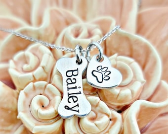 May include: A silver necklace with a bone-shaped pendant engraved with the name "Bailey" and a heart-shaped pendant with a paw print. The pendants hang from a delicate chain.