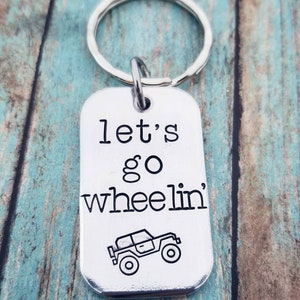 Puede incluir: Llavero de plata con una etiqueta que dice "let's go wheelin'" y una pequeña ilustración en blanco y negro de un jeep.
