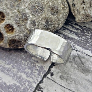 May include: A silver hammered metal ring with a wide band. The ring is open and has a slightly curved shape.
