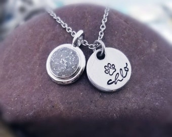 May include: Silver necklace with two charms. One charm is a round silver pendant with a paw print and the name "Chloe" engraved on it. The other charm is a round silver pendant filled with white glitter.