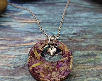 Op de afbeelding: Een zilveren ketting met een ronde hanger gemaakt van helder hars met gedroogde bloemen en een klein zilveren hartje.
