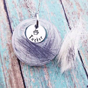 May include: A silver necklace featuring a circular pendant filled with grey fur. A smaller silver charm displays a paw print and the name "Tucker". The backdrop is a weathered, turquoise-coloured wooden surface.