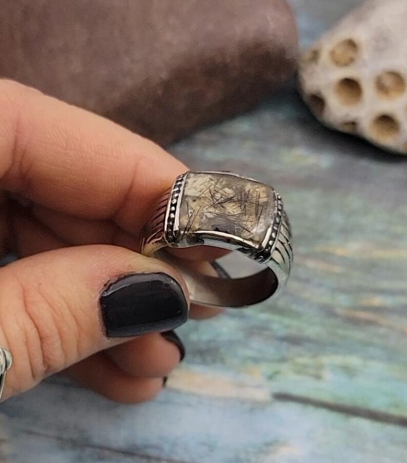 May include: A silver ring with a square-shaped stone set in the center. The stone is clear with black and brown inclusions. The ring has a textured band.