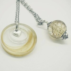 May include: A silver necklace with two pendants. One is a clear, circular resin pendant with blonde hair and a small silver heart charm. The other is a clear sphere with hair inside. Both pendants hang from a silver chain.