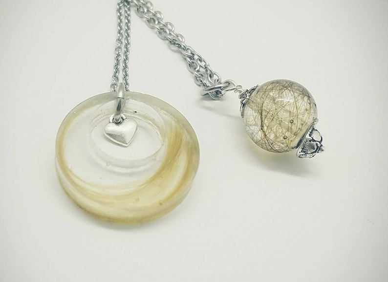 May include: A silver necklace with two pendants. One is a clear, circular resin pendant with blonde hair and a small silver heart charm. The other is a clear sphere with hair inside. Both pendants hang from a silver chain.